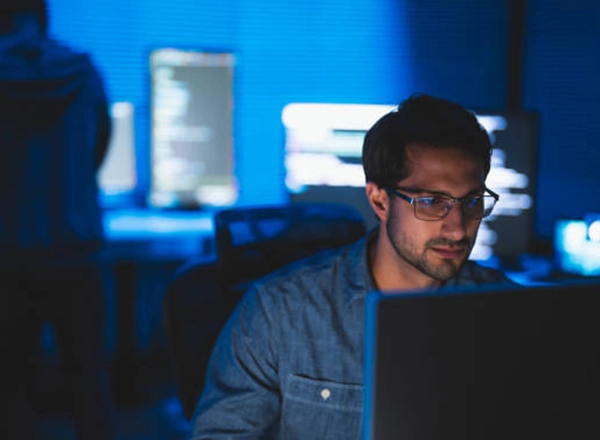 A focused male software developer wearing glasses is seated in a dark office, looking at a computer screen. In the background, another developer is also working in the dimly lit space.