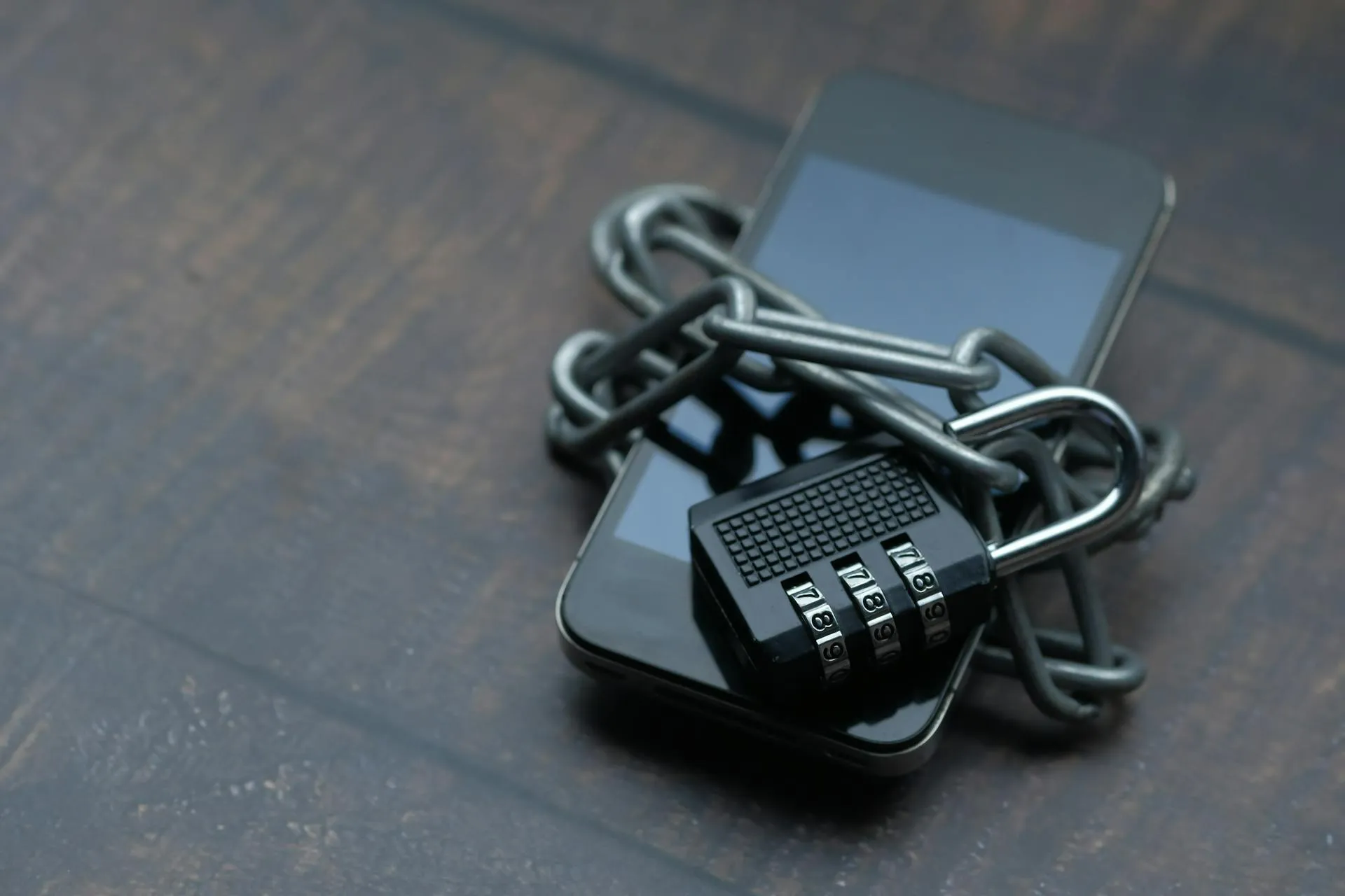 A black smartphone wrapped in a silver chain with a numeric combination padlock resting on top, set against a dark brown surface. The image represents locked-down mobile security, illustrating the need for multi-factor authentication and passkey protection against phishing attacks for Houston businesses.