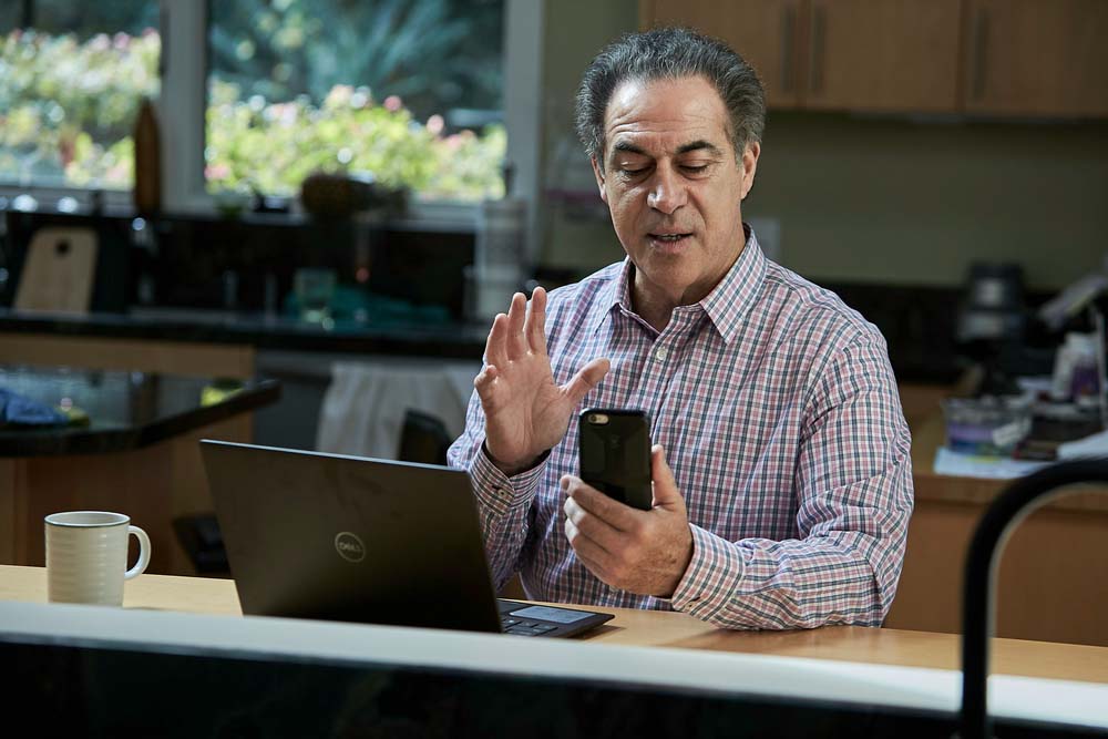 A financial professional using Elevate Technology's mobile app to make a video call from his kitchen. He is seated at a counter with a laptop and a mug, holding a smartphone and waving, demonstrating the flexibility of a remote work solution.