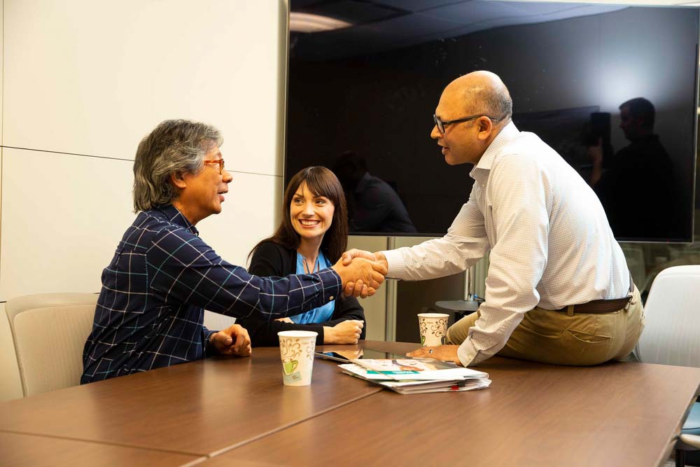 A business meeting in a legal office where three people are seated at a table. Two men are shaking hands, showcasing client or team collaboration potentially enhanced by Elevate Technology.