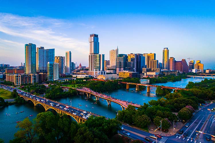 An aerial shot of the Austin, Texas skyline at sunset, showing the downtown buildings lit by the golden hour light, the Colorado River flowing through the city with bridges, and traffic moving on the roadways. This image is for Elevate Technology.