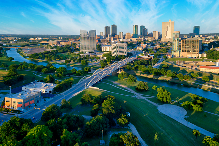 An aerial view of the Fort Worth, Texas skyline and the Trinity River, with a modern white arched bridge crossing the water and lush green parks along the banks. This image is for Elevate Technology.
