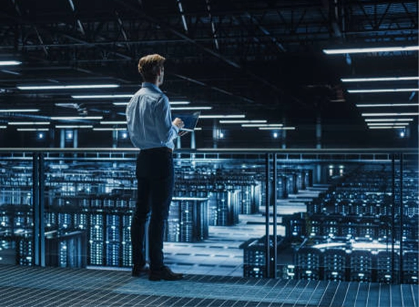 A professional man holding a tablet, standing on a platform and looking out over the rows of servers in a modern data center.