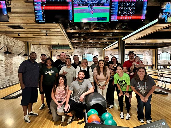 A large group of Elevation Technology employees smiling and posing for a group photo during a company bowling event in 2024. They are standing in the bowling alley, with a large bowling ball and pins in the foreground and screens displaying a game in the background.