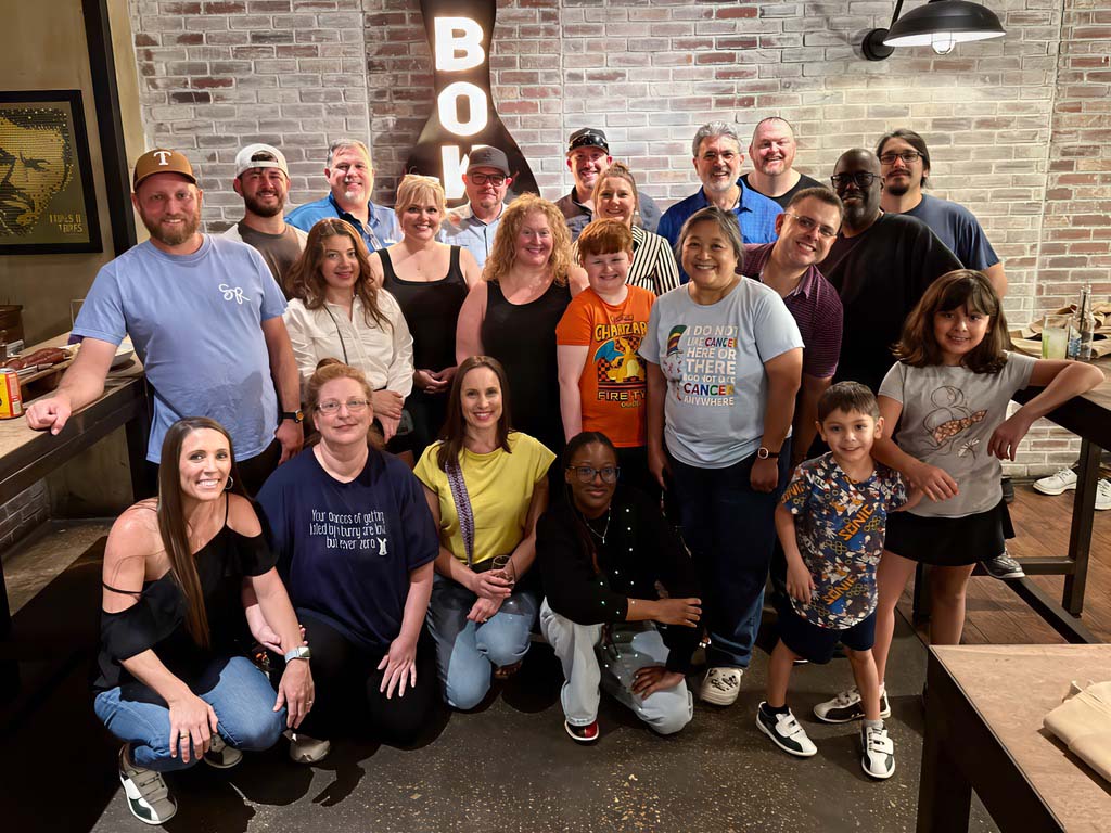 Elevate Technology photo of a large group of smiling adults and children posing together indoors in front of a brick wall, with a partial 
