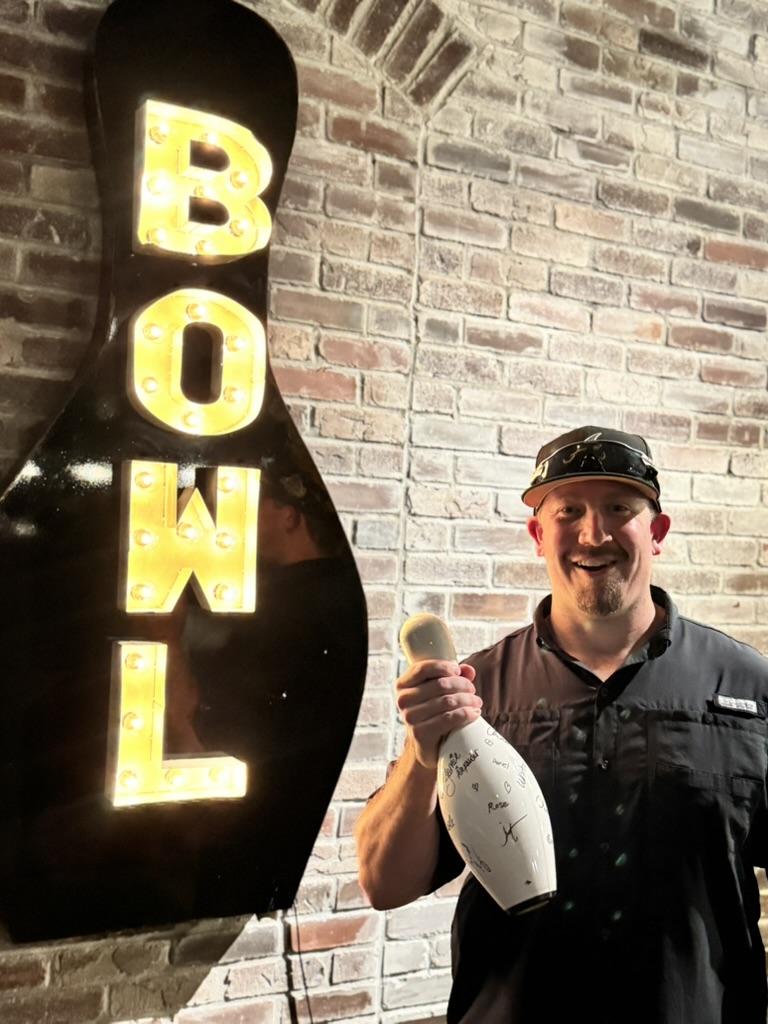 Elevate Technology photo of a smiling man in a black shirt and baseball cap holding a signed white bowling pin, standing next to a large, illuminated 