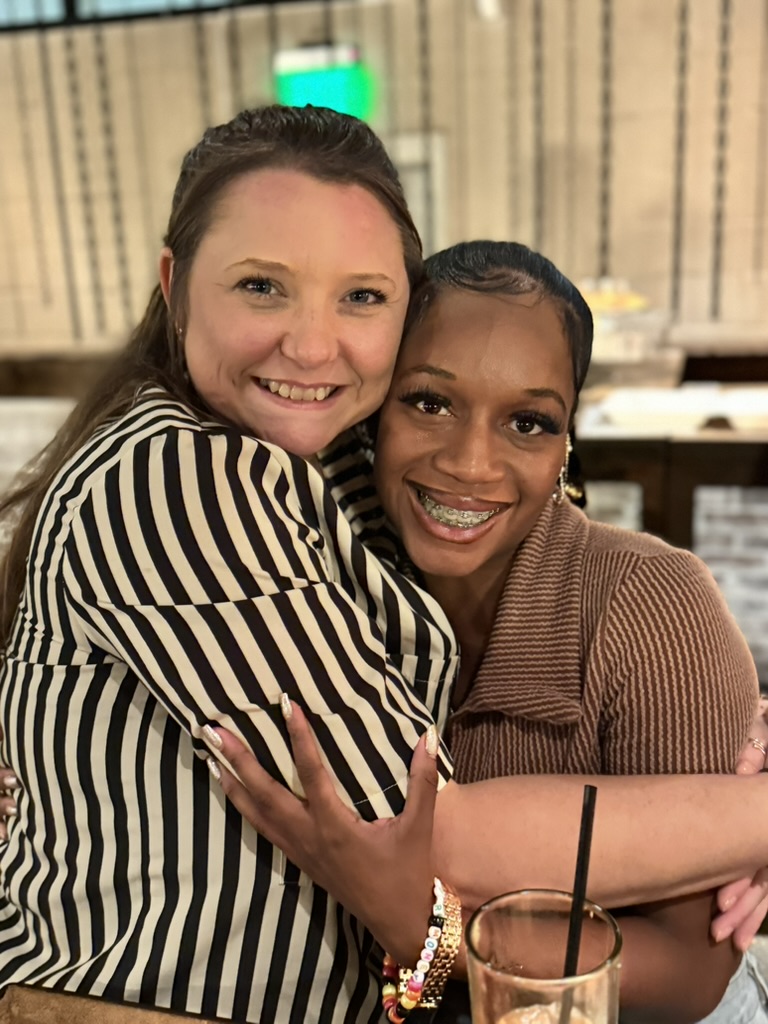 Elevate Technology close-up photo of two smiling women, Nicole and Danaja, embracing one another at a social event. One woman is wearing a black and white striped shirt, and the other is wearing a brown ribbed shirt.