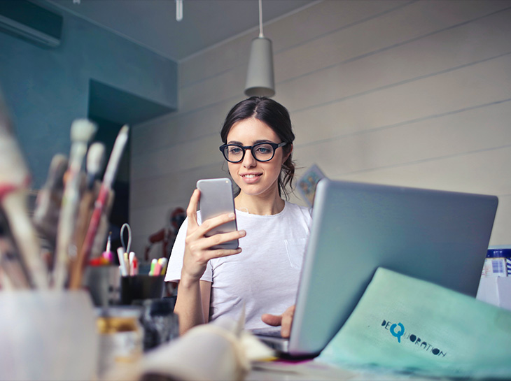 An Elevate Technology remote employee wearing glasses, looking at her smartphone while working on a laptop at a desk.