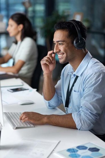  Elevate Technology image of a smiling, dark-haired man in a headset sitting at a desk, typing on a keyboard, with a female colleague visible in the background, representing a managed IT service desk.