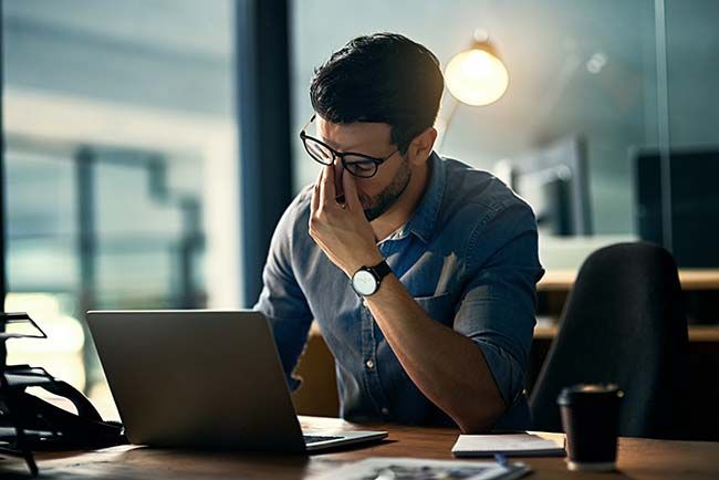 A powerful stock photo for Elevate Technology's managed IT services, showing a man who is an office worker looking stressed and rubbing his eyes while sitting in front of his laptop at his desk. 
