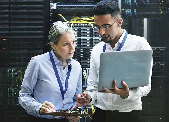 A professional stock photo for Elevate Technology's data center services. A male and a female IT professional are working together in a server room, with racks of servers visible in the background. The man holds a laptop and points to something on the screen while the woman looks on with a focused expression, representing teamwork and expertise in managing complex IT infrastructure.      Analysis