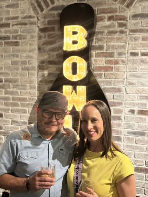 Elevate Technology photo of a smiling man, Alfredo, and a smiling woman posing together, each holding a glass, in front of a brick wall and a large, illuminated "BOWL" sign. The man is wearing a cap and glasses, and the woman is wearing a yellow shirt.
