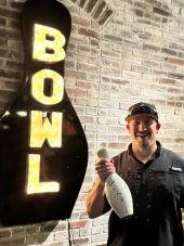Elevate Technology photo of a smiling man in a black shirt and baseball cap holding a signed white bowling pin, standing next to a large, illuminated "BOWL" sign shaped like a black bowling pin against a brick wall.