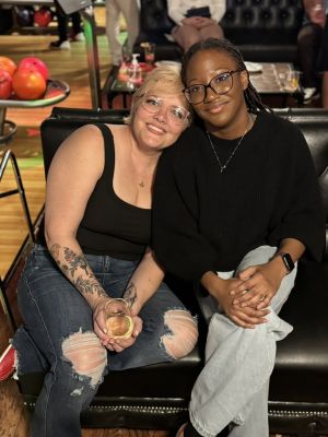 Elevate Technology photo of two women, including Angela (right), seated together and smiling at a social event. The woman on the left has her head resting on the other's shoulder, holds a wine glass, and wears a black tank top and ripped jeans. The woman on the right wears a black sweater and light jeans. Bowling balls and pins are visible in the background.