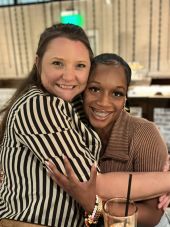 Elevate Technology close-up photo of two smiling women, Nicole and Danaja, embracing one another at a social event. One woman is wearing a black and white striped shirt, and the other is wearing a brown ribbed shirt.