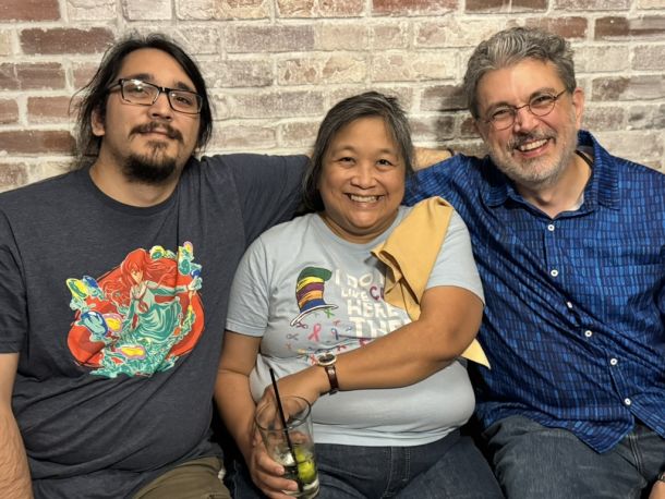 Elevate Technology photo of three smiling people, including Chris (center), with her arms around a man on the left wearing glasses and a graphic t-shirt, and another man on the right wearing a blue patterned shirt, all posing in front of a brick wall.