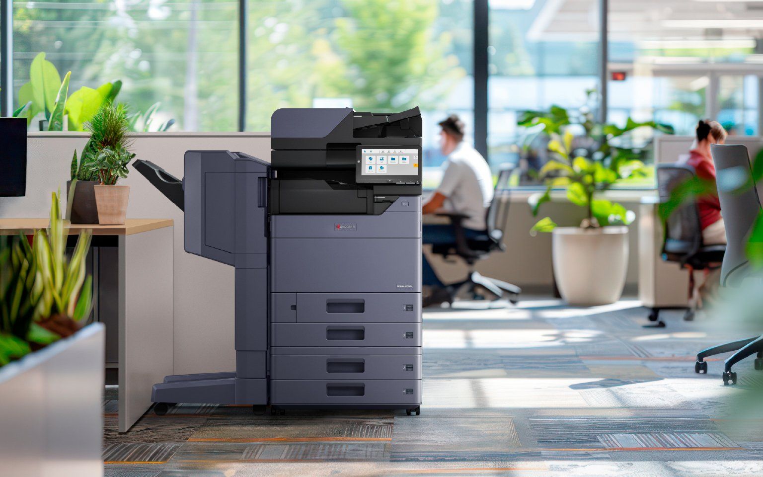 Elevate Technology photo showing a black Kyocera multi-function printer/copier in a modern office environment with plants and a blurred person sitting at a desk in the background.