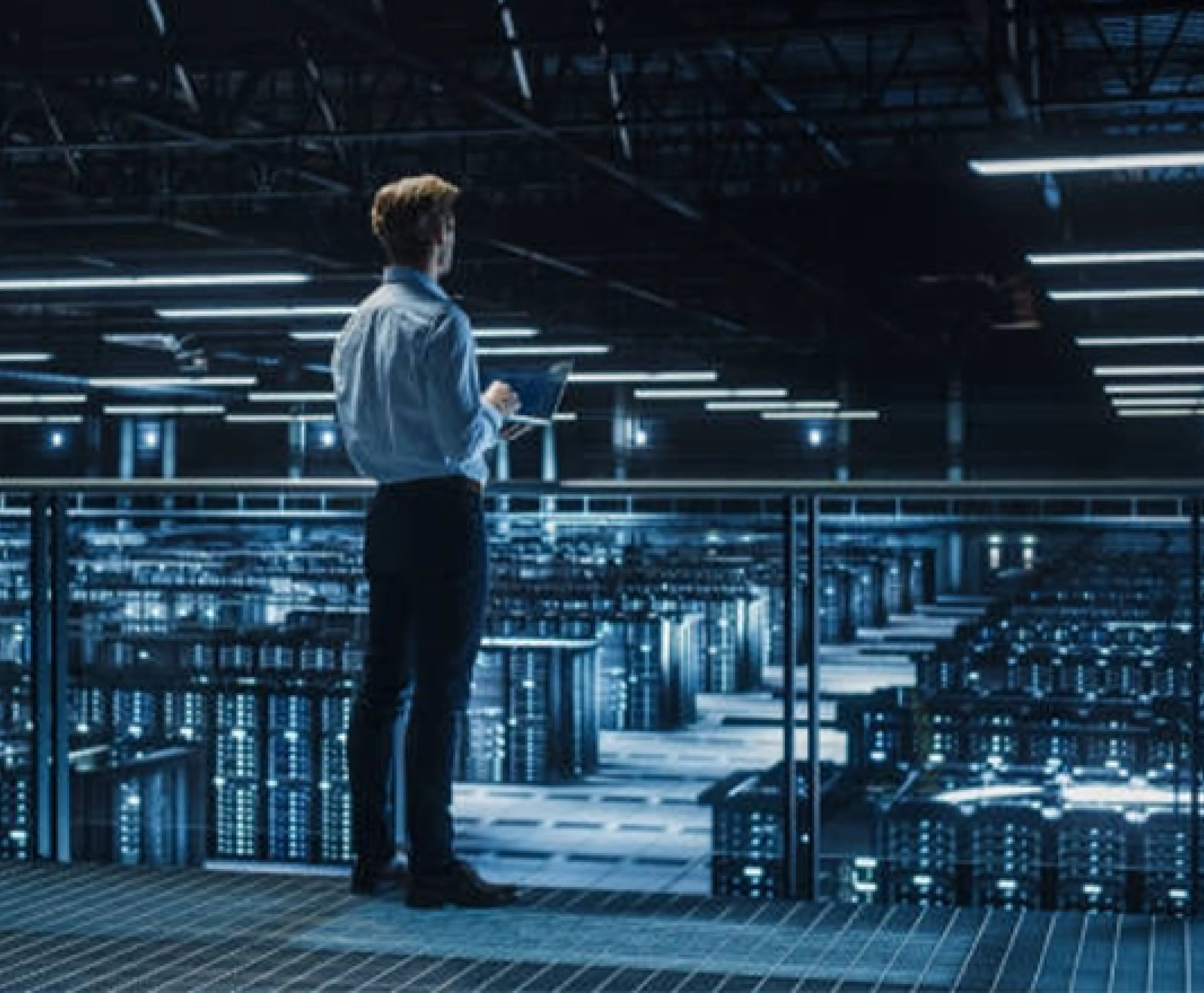 A professional man holding a tablet, standing on a platform and looking out over the rows of servers in a modern data center.