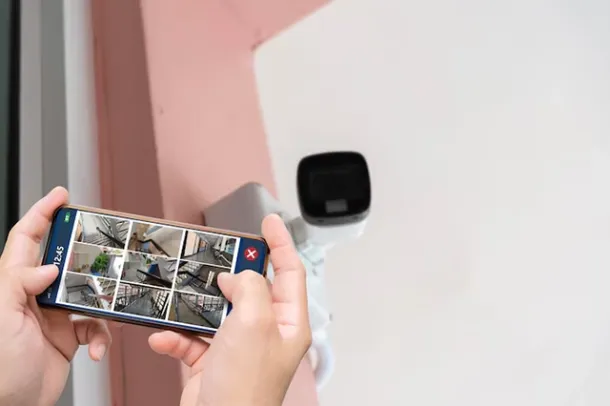 Elevate Technology person holding a smartphone displaying a grid of security camera feeds in front of an outdoor wall-mounted security camera.