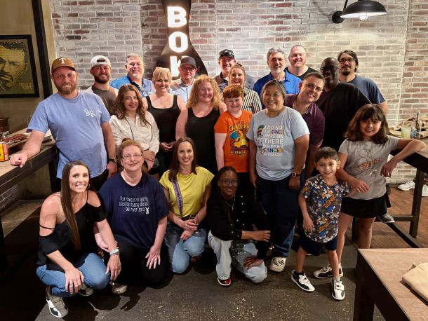 Elevate Technology photo of a large group of smiling adults and children posing together indoors in front of a brick wall, with a partial "BOWL" sign visible overhead, likely taken during a team bowling night event in 2025.